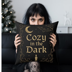 May include: Black square pillow with gold text and accents. The pillow features the words "Cozy in the Dark" with a crescent moon, bat, and candle design. The pillow is held up by a person.