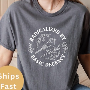 Op de afbeelding: Donkergrijs t-shirt met de tekst "RADICALIZED BY BASIC DECENCY" in een wit cirkelvormig ontwerp met een vogel op een tak. Het shirt is een comfortabele, casual stijl.