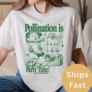 May include: A light gray t-shirt with a green graphic that reads "Pollination is Party Time!" The graphic includes illustrations of flowers, bees, and scientific diagrams. The shirt is made of a soft material.