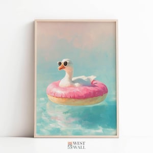 May include: A framed wall art print featuring a white goose wearing black sunglasses, floating in blue water on a bright pink donut-shaped pool float. The bottom of the print displays the text West & Wall in a small, dark grey font.