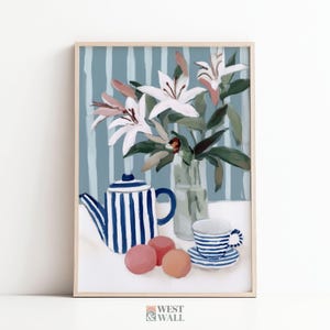 Könnte beinhalten: Gerahmter Kunstdruck mit einem Stillleben mit einer blau-weiß gestreiften Teekanne, Tasse und Untertasse. Eine Vase mit weißen Lilien mit rosa Akzenten befindet sich hinter dem Teeservice. Drei rosa und orangefarbene Kugeln befinden sich vor der Teekanne.