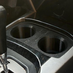 以下が含まれることがあります： 2つの円形開口部を備えた黒いプラスチック製の車のカップホルダー。 カップホルダーは、ギアシフトの隣にある車のコンソールに統合されています。 コンソールは木目模様とシルバーのトリムを備えています。