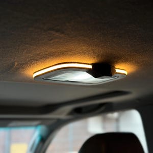 Puede incluir: Una luz interior rectangular de vehículo montada en un techo de tela gris oscuro. La lámpara cuenta con una lente blanca translúcida rodeada por un borde LED ámbar brillante y un componente de carcasa de plástico negro en un lado.