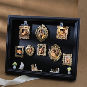 Może przedstawiać: Czarna gablota z miniaturowymi zdjęciami kotów i rodziny w złotych ramkach. Małe figurki kotów i miniaturowe kompozycje kwiatowe umieszczone są pod ramkami. Biała wstążka znajduje się na pierwszym planie.