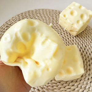 Peut inclure: Gros plan sur trois objets en forme de fromage. Le plus grand est de forme arrondie, les deux autres sont en forme de cube. Tous sont de couleur jaune pâle avec des trous, ressemblant à du fromage suisse. Les objets sont sur une surface tissée brun clair.