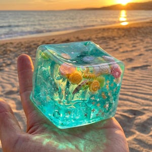 May include: A clear, square resin paperweight with a turquoise hue, encasing various seashells and gold star-shaped glitter. The paperweight is held in a hand, with a beach and sunset in the background.