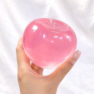 May include: A pink glass apple with a clear stem. The apple is held in a hand, and the background is a white, textured fabric. The apple has a glossy finish and a rounded shape.