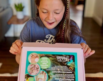 May include: A young girl smiles while holding a pink box. The box features a teal and white design with the words "Welcome Luna Sky" and "My Real Fairies." The box also has images of fairy gardens.