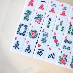 May include: A collection of white rectangular mahjong tiles arranged in a grid. The tiles feature various engraved symbols, including Chinese characters, numbers, directional letters E, S, W, N, and colorful illustrations of birds, flowers, and circles.