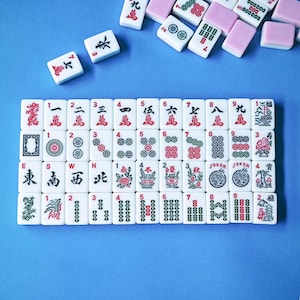 May include: A collection of white rectangular mahjong tiles featuring black, red, and green engraved symbols, numbers, and characters. The tiles are arranged in a grid on a blue surface, with several loose tiles scattered near the top edge.