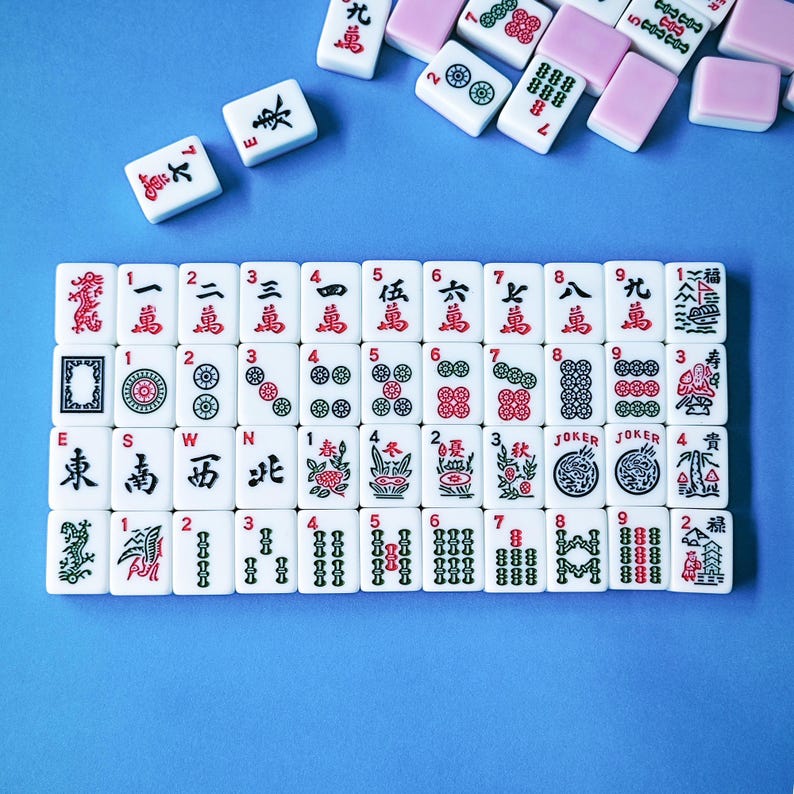 May include: A collection of white rectangular mahjong tiles featuring black, red, and green engraved symbols, numbers, and characters. The tiles are arranged in a grid on a blue surface, with several loose tiles scattered near the top edge.
