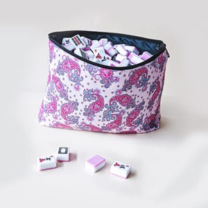 May include: A zippered fabric pouch with a light pink background and a repeating dark pink and blue paisley pattern, filled with white and pink mahjong tiles. Four additional mahjong tiles are placed in front of the pouch on a plain surface.