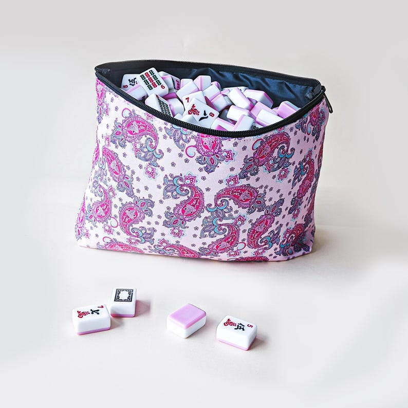 May include: A zippered fabric pouch with a light pink background and a repeating dark pink and blue paisley pattern, filled with white and pink mahjong tiles. Four additional mahjong tiles are placed in front of the pouch on a plain surface.