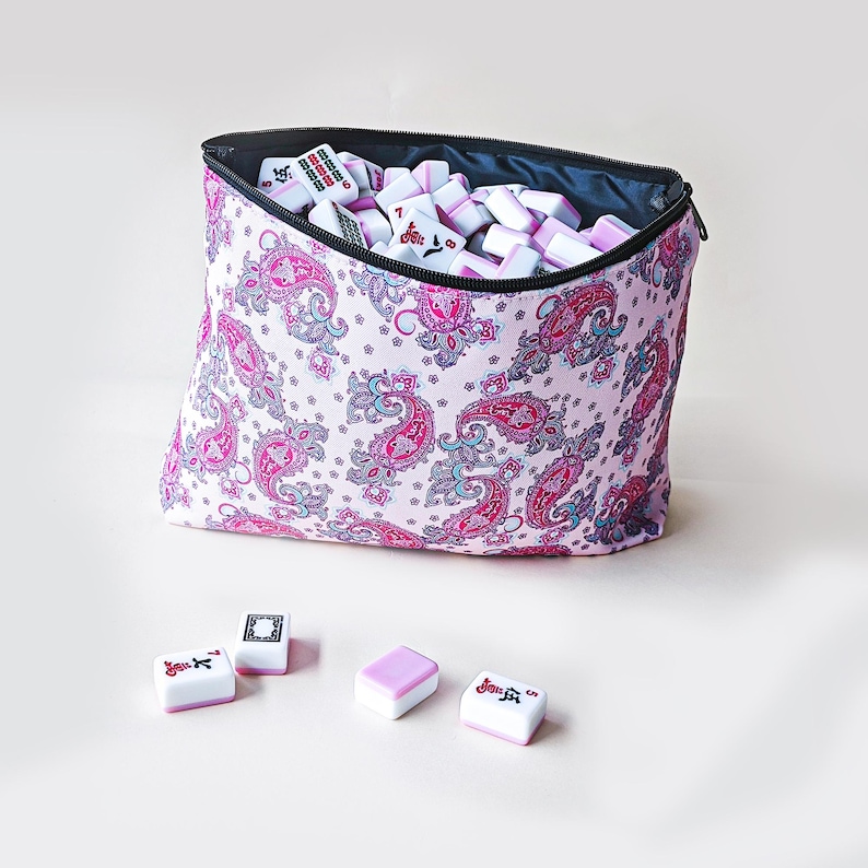 May include: A zippered fabric pouch with a light pink background and a repeating dark pink and blue paisley pattern, filled with white and pink mahjong tiles. Four additional mahjong tiles are placed in front of the pouch on a plain surface.