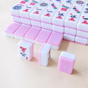 May include: A collection of rectangular Mahjong tiles featuring a white top section and a pastel pink bottom section. The tiles are arranged in rows, with some displaying red and black Chinese characters, numbers, and circular floral patterns.