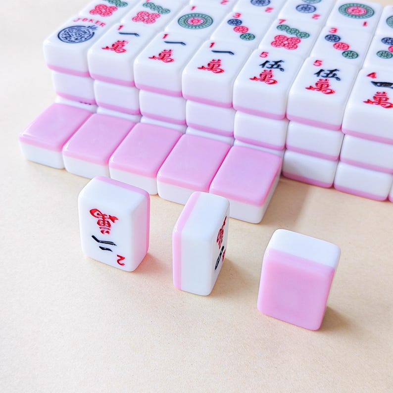 May include: A collection of rectangular Mahjong tiles featuring a white top section and a pastel pink bottom section. The tiles are arranged in rows, with some displaying red and black Chinese characters, numbers, and circular floral patterns.