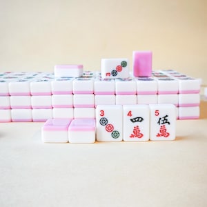 May include: A set of rectangular mahjong tiles with white faces and pink backs. The tiles are arranged in rows and stacks. Some tiles feature red and green circular symbols, while others display the numbers 3, 4, and 5 alongside Chinese characters.