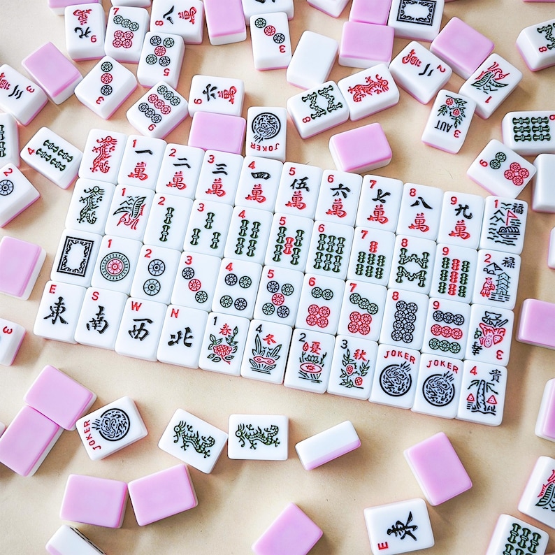 May include: A collection of white mahjong tiles featuring red, green, and black ink designs, including Chinese characters, numbers, and symbols. A central rectangular grid of tiles is surrounded by scattered tiles and plain pink-backed tiles.