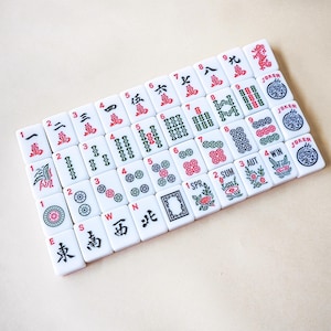 May include: A collection of white rectangular mahjong tiles arranged in a grid. The tiles feature various red, green, and black engraved symbols, including Chinese characters, numeric values, seasonal icons, and joker labels with dragon illustrations.