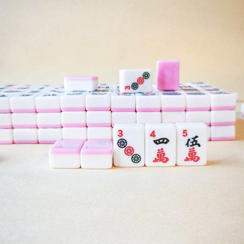 May include: A set of rectangular mahjong tiles with white faces and pink backs. The tiles are arranged in rows and stacks. Some tiles feature red and green circular symbols, while others display the numbers 3, 4, and 5 alongside Chinese characters.