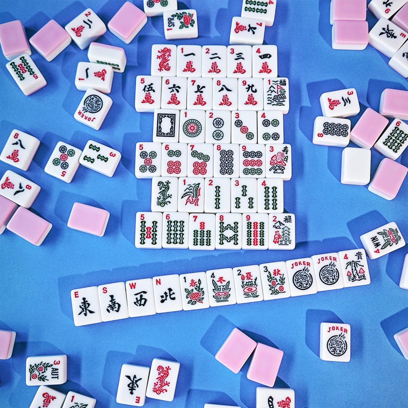 May include: A collection of white Mahjong tiles with red, green, and black ink designs, including Chinese characters, numbers, and symbols. The tiles are arranged in a central grid and a horizontal row on a blue surface, with scattered tiles nearby.