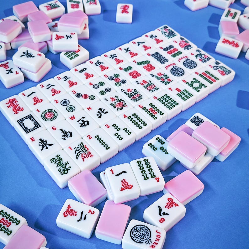 May include: A set of white Mahjong tiles with pink backs, featuring various red, green, and black engraved symbols, characters, and numbers. The tiles are arranged in a rectangular grid formation with additional loose tiles scattered around.