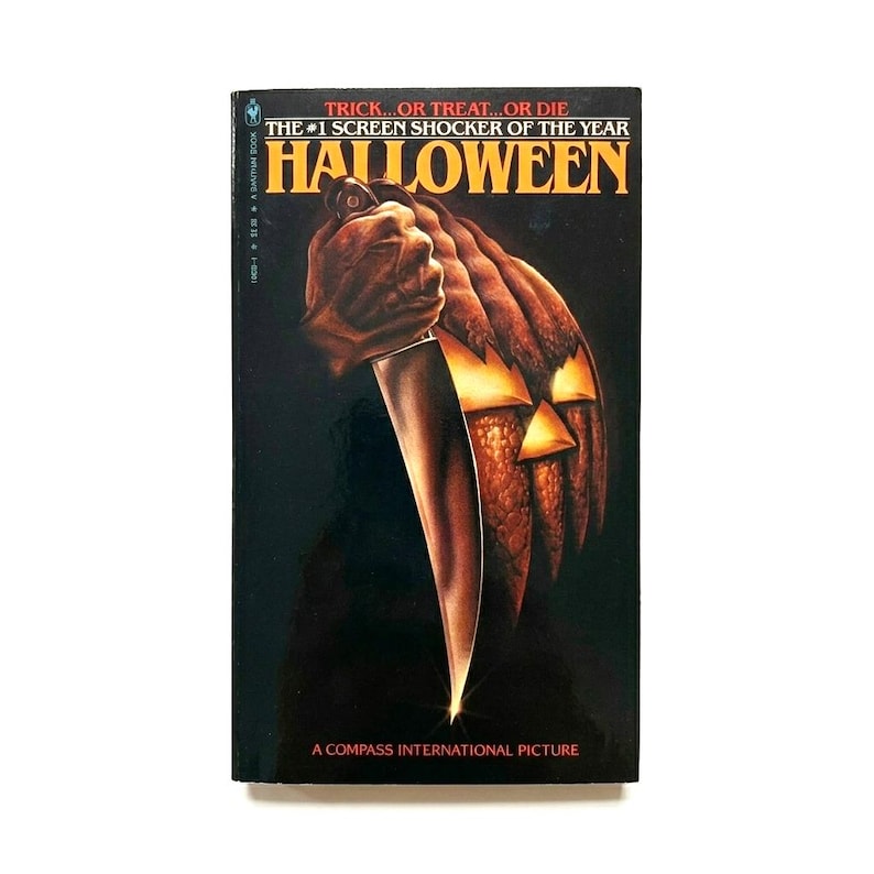 May include: A vintage book cover for the horror film "Halloween." The cover shows a hand holding a knife, with a carved pumpkin in the background. The title "Halloween" is in large orange letters.
