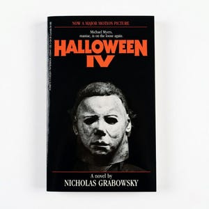 May include: A paperback book with a black cover featuring the title "HALLOWEEN IV" in orange. The cover shows a white mask and the text "NOW A MAJOR MOTION PICTURE". The author's name, NICHOLAS GRABOWSKY, is also visible.