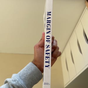 Puede incluir: Lomo de libro blanco con el t&iacute;tulo "MARGIN OF SAFETY" en letras may&uacute;sculas azules. El libro se sostiene verticalmente, con el nombre del autor y la editorial en la parte inferior. Un libro de negocios.