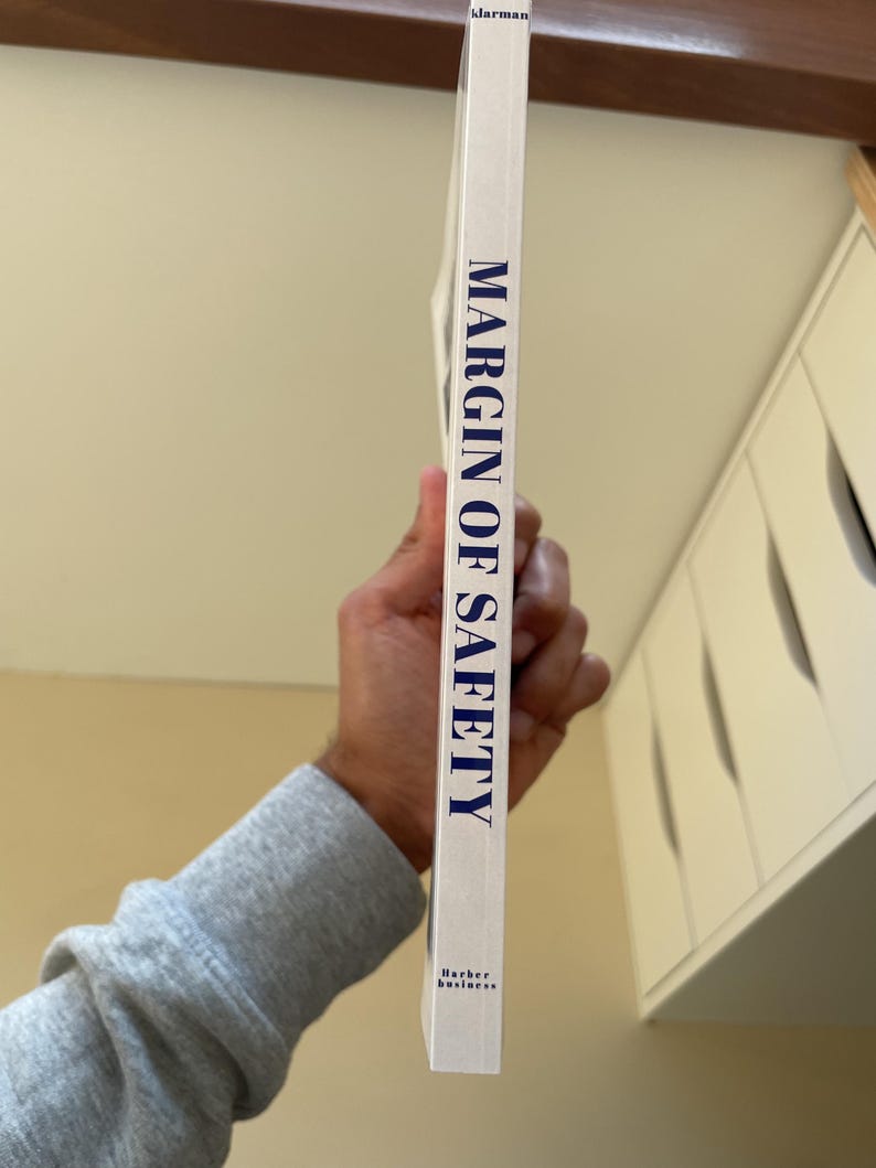 Puede incluir: Lomo de libro blanco con el t&iacute;tulo "MARGIN OF SAFETY" en letras may&uacute;sculas azules. El libro se sostiene verticalmente, con el nombre del autor y la editorial en la parte inferior. Un libro de negocios.