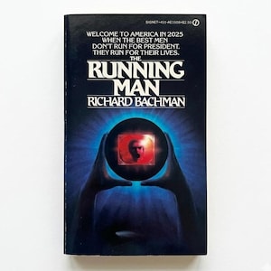 Puede incluir: Un libro de bolsillo vintage titulado "The Running Man" de Richard Bachman. La portada es azul oscuro con texto blanco, incluyendo el título y el nombre del autor. Un gráfico muestra la cara de una persona en un orbe brillante sostenido por manos. El texto dice "Welcome to America in 2025".