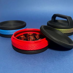 May include: Several dice containers in different colours, including red, blue, and black. The containers have a textured design and are open, revealing dice inside. One container has a handle.