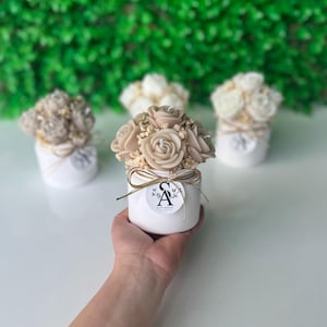 Op de afbeelding: Een beige roosvormige kaars in een witte cilindrische houder, versierd met kleine gedroogde bloemen en een gouden lint. Een rond label met de letters SA en de tekst GIFT HOUSE is aan de voorzijde bevestigd. Twee soortgelijke kaarsen zijn erachter.