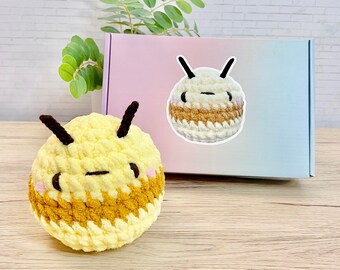 Peut inclure: Deux peluches d'abeilles en crochet, l'une jaune et marron, l'autre blanche et marron, avec des antennes noires et des visages souriants. L'abeille jaune et marron est posée sur une surface en bois, tandis que l'abeille blanche et marron est sur une boîte rose et bleue.
