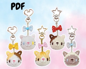 May include: Five crocheted cat head keychains with different colored bows and key rings. The keychains are white with brown, gray, or orange accents. The bows are yellow, blue, pink, red, and light blue.
