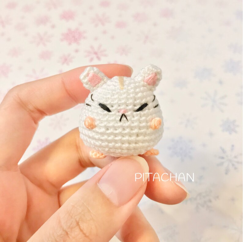 May include: White crocheted hamster amigurumi with pink ears and a grumpy expression. The hamster is being held in a hand. The background is a white and pink snowflake pattern.
