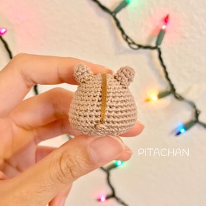 May include: A small crocheted brown bear with a gold thread down the back. The bear is being held by a hand.