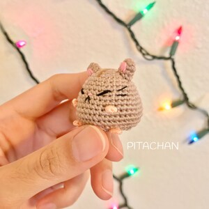 May include: A small crocheted hamster with brown fur, pink ears, and black eyes. The hamster is winking and has a pink nose. The hamster is being held in a hand against a background of colorful Christmas lights.