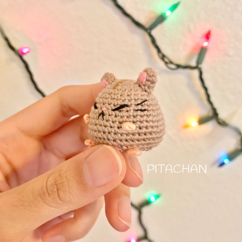 May include: A small crocheted hamster with brown fur, pink ears, and black eyes. The hamster is winking and has a pink nose. The hamster is being held in a hand against a background of colorful Christmas lights.
