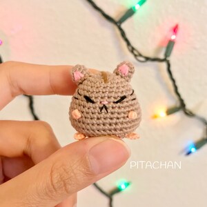 May include: A small crocheted hamster with brown fur, pink ears, and black eyes. The hamster has a grumpy expression and is holding its paws up.