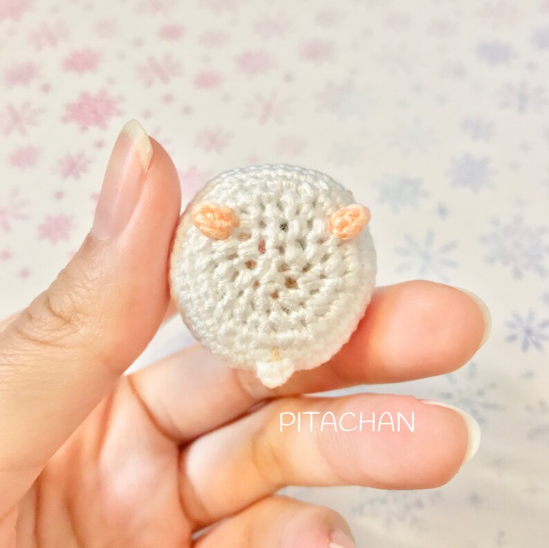 May include: A small white crocheted hamster with peach-colored ears. The hamster is being held in a hand. The background is a white and pink snowflake pattern.