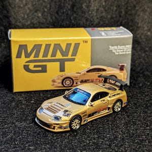 May include: A gold-colored diecast model car of a Toyota Supra (A80) Top Secret GT-300. The model car has black accents, a rear spoiler, and blue tinted windows. The packaging box is yellow with the words "MINI GT" printed on it.