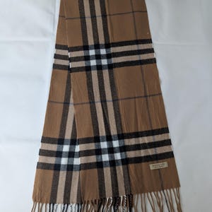 Könnte beinhalten: Ein beigefarbener Schal mit einem klassischen Karomuster aus schwarzen, weißen und roten Streifen. Das rechteckige Accessoire hat ein kleines rechteckiges Etikett in der unteren Ecke mit dem Text Burberry London England und Fransen an beiden Enden.