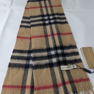 Könnte beinhalten: Ein beigefarbener Schal mit dem ikonischen Burberry-Karomuster in schwarzen, weißen und roten Streifen. Der Schal hat eine weiche, gebürstete Textur und gefranste Enden. Ein weißes Stoffetikett mit der Aufschrift Burberry ist angebracht.