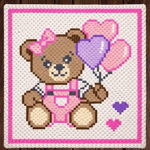 May include: A crocheted square with a brown teddy bear wearing pink dungarees and a bow, holding heart-shaped balloons. The design has a pink border and heart accents, set against a white background.