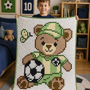 May include: A white and green crocheted blanket featuring a soccer-themed teddy bear design. The bear wears a green and yellow soccer uniform and holds a black and white soccer ball. The blanket has a green border.