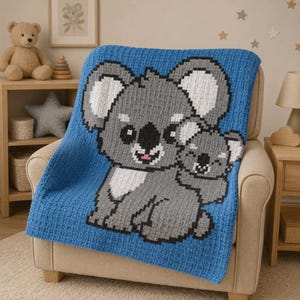 Puede incluir: Una manta azul tejida con un diseño de un koala gris y su bebé. Los koalas tienen ojos y narices negros y bocas rosas. La manta está sobre un sillón beige, creando una escena acogedora y acogedora.