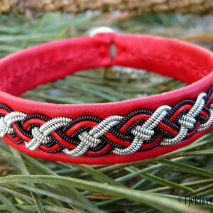 May include: Red leather bracelet with a silver and black braided design. The bracelet is on a bed of green pine needles.
