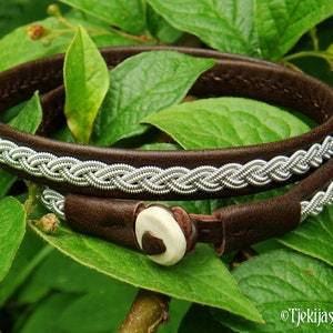 Könnte beinhalten: Ein braunes Lederarmband mit einem geflochtenen Silberdesign und einem Knochenverschluss. Das Armband liegt auf einem grünen Blatt-Hintergrund.