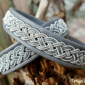 Könnte beinhalten: Zwei graue Lederarmbänder mit gewebten Designs aus Silberdraht. Die Armbänder sind übereinander gestapelt.